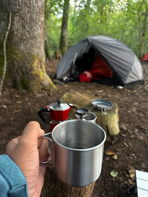 Tasse en métal au premier plan, cafetière rouge et tente installée parmi les arbres.